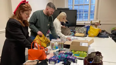 Four people, a woman with ginger hair, a man with brown hair, a woman with blonde hair and a woman with black hair are standing over a table. The table has boxes of sanitary pads, gloves, body spray and handbags. They are packing the handbags with the items.