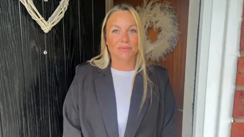 A woman with long blonde hair, a grey jacket and white top stands in the doorway of a house.