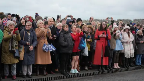 PA Media Members of the public wait for the arrival of the Princess Royal to officially open the Gull Wing Bridge in Lowestoft, Suffolk. They are standing on a pavement on the edge of the bridge and many of them are taking pictures of the princess on their phones, while others are applauding