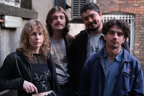 BBC Four members of Brown Horse standing together and looking at the camera outdoors in front of brick and concrete walls.