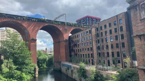 Photograph of the brick railway viaduct in Stockport town centre. The image shows the River Mersey and a cotton mill underneath one of the arches. 