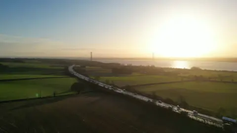 BBC A drone image of East Yorkshire. The sun is rising over the Humber Bridge which visible in the distance along with the Humber Estuary. Cars and lorries are visible on the A63 road which is running across the image and flanked by green fields.