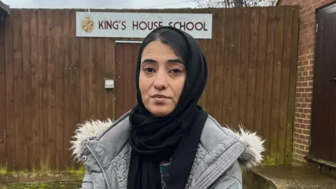 A woman wearing a black hijab and grey coat standing in front of a wooden gate which has a sign which reads "King's House School". There is a red brick building to the right.