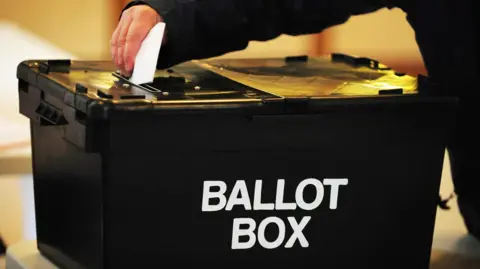 PA Media A black box with the words 'Ballot Box' written on the front in white. A hand is above placing a white piece of paper in a hole on the top. 