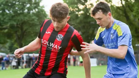 David McDonald A footballer with red hair and a beard with a red and black striped top shields the ball from a man in a blue shirt with gold stripes on it
