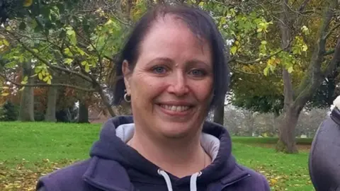 A woman with dark hair and a purple top standing on a grassy area with trees behind her