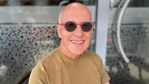 BBC A man sat outside a cafe, wearing a brown t shirt. He has a bald head and blue a red sunglasses. He is smiling at the camera