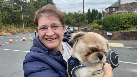 A woman in a navy puffer jacket and grey jumper smiles as she holds up a small dog in the street, with traffic cones across the road entrance.