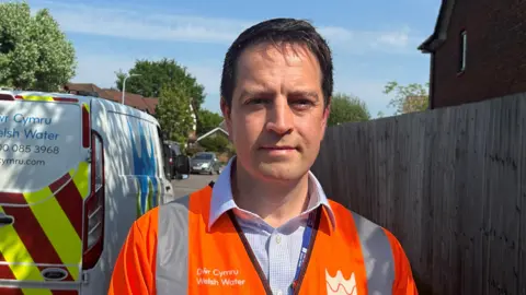 Chris in a Welsh Water orange high vis vest looks into camera, with a Welsh Water van in the background. He has short dark hair and is wearing a blue and white chequered shirt underneath the vest.