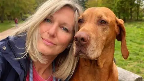 Family handout Sam Carter is wearing a red t-shirt and navy blue jacket sitting next to her dog in a park