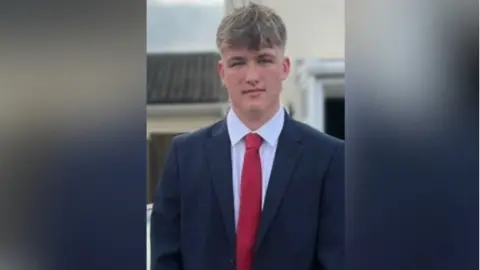 Joshua Blake wearing a suit and a red tie. He is standing in front of a building and looking at the camera.
