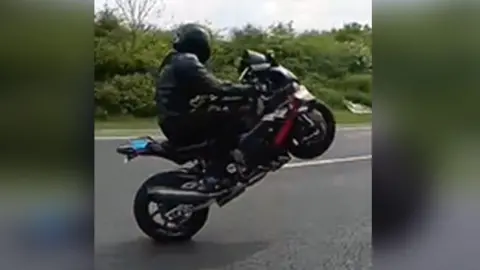 Essex Police A motorcyclist on his machine wearing black leather and a black helmet. The bike they are on is also black and the front wheel is off the ground as they perform a "wheelie".