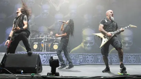 Three members of Slayer, Frank Bello, Joey Belladonna and Scott Ian playing guitars and singing on stage. There is a set of drums in the background with pictures of skulls wearing crowns serving as a backdrop on the stage. 