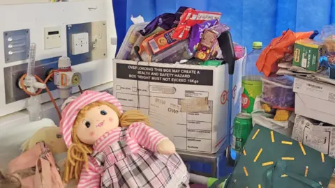 With two cloth dolls, a blonde woolen hair and a striped pink hat, lie on the bed of Jessie's hospital next to the general hospital tube and oxygen outlet. The beverages and cardboard boxes on the bedside table are high stacks with biscuits, snacks, art materials and other property. 