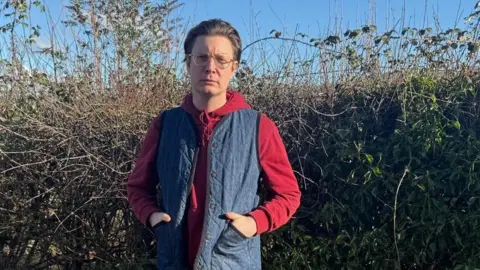 Handout Hofmann standing outdoors on a paved path in front of a tall, dense hedge made up of bare branches and green foliage. He is wearing a red hoodie underneath a blue sleeveless padded vest, along with light grey jogger-style trousers and dark trainers. His hands are tucked into the pockets of the vest. The sky above is bright and clear.