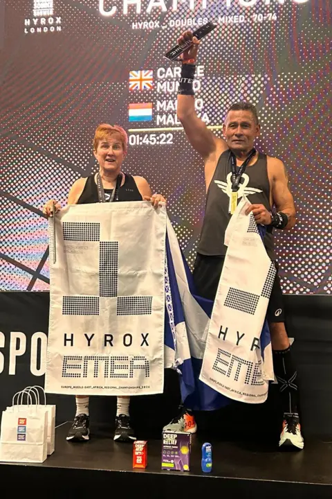 Stuart Amory Carole Munro and Jim Malinka stand on the podium holding white and black Hyrox flags.