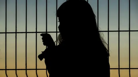 Young girl looks through metal fence, at sunset. She is silhouetted. 