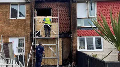 Julie Smith/BBC The outside of the house which now has scaffolding in front of it with two men putting up the cardboard.