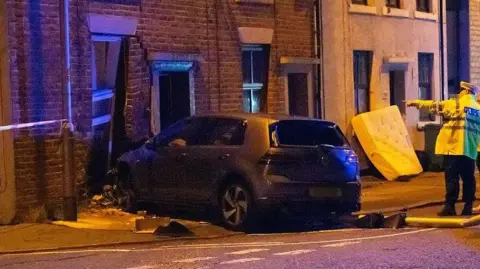 Silver VW car has smashed into a terraced house which has destroyed the front  window and door. Debris is strewn across the road including a lamppost which has been flattened and is on the road. Police tape has cordoned the area off. 