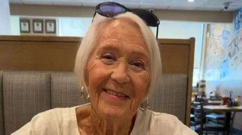 Gloria Stephenson smiling into the camera. She has short, white hair and is wearing light pink lipstick, gold hoop earrings and a white top. She looks to be sitting in a restaurant.