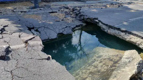 Thames Water A close up of a road with a large hole filled with water and surrounded by cracked and broken tarmac.