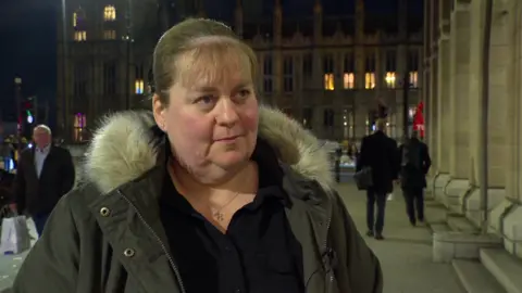 A woman with brown hair that is tied up stands looking away from the camera. She is wearing a parka coat with faux fur lining the hood. She has a black top on underneath.