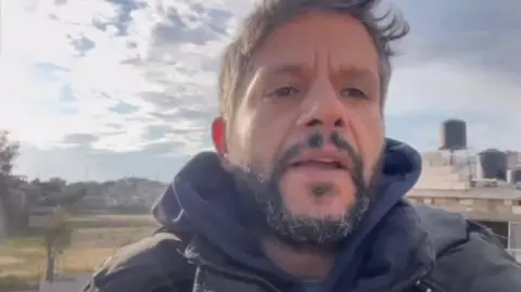 EPA Husam al-Masri, bearded, wearing a blue hoodie under a dark jacket, shown with buildings in the distance behind him, one of which has two large water containers on the roof