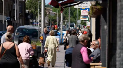 A busy shopping street with numerous people sitting at pavement cafes