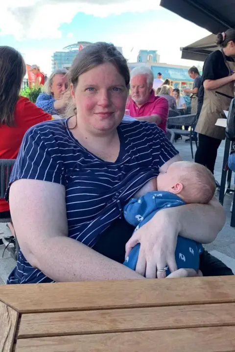 Louise Owen Louise is sat at an outdoor cafe breastfeeding. There are other customers in the background.