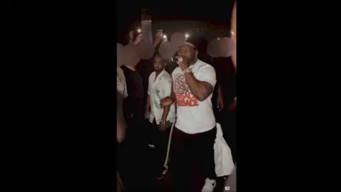 Supplied Hassan, wearing a silky button up shirt, stands next to 50 Cent, who is holding a microphone.