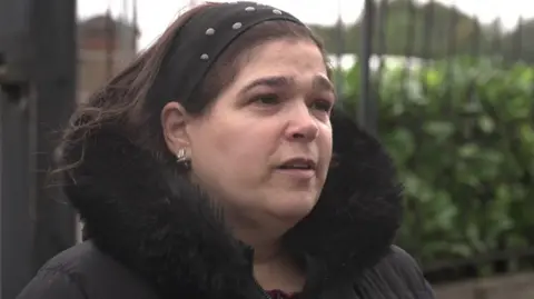 Hannah Salomon speaks during an interview. She wears a dark hairband and wears a dark coat as she stands outside the cemetery railings.