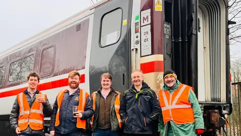 Five members of the 225 Preservation CIC team smile at the camera in front of one of their train passenger carriages. They all wear orange high-vis jackets and some of them hold cups of drinks. 