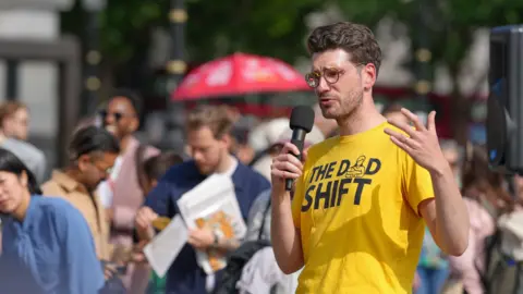 The Dad Shift Alex is wearing a yellow t-shirt with The Dad Shift in black writing on it. He is holding a microphone as he makes a speech at a protest rally in the UK.