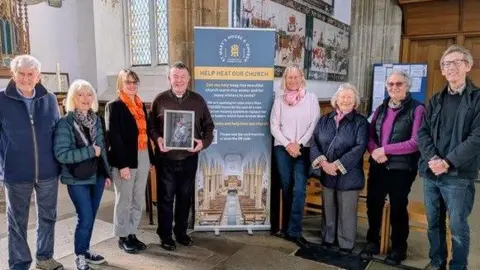 Contributed A group of people associated with St Mary's Church stand together in a group and smile at the camera. In the middle of them is a tall information board about their heating fund. One of the men, the Reverend Nigel Prior, holds the Maggi Hambling painting in his hands. 