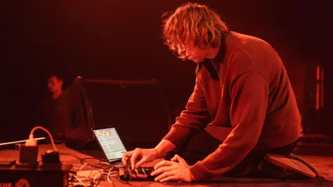 Daniel Avery is sitting on a stage while using DJ equipment. He is wearing a jumper and has shoulder length, blonde hair.