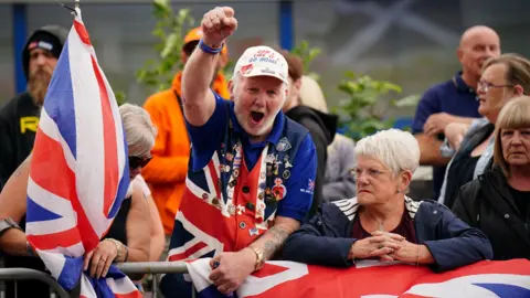 A group of PA media stands at a metal barrier. A woman wearing sunglasses holds a union flag. There is a man in the middle of the scene wearing Kamarkot in the colors of the Sangh flag. The Vascut Pin is covered in the badge. The man, who is wearing a hat, is raised with a hand near it because he shouts. A woman next to her is looking at the distance, bending over the flag wrapped over the barrier.