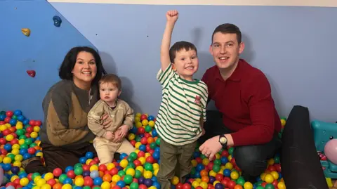a family of four - a woman, man and two young boys - in a ball pit. The older boy, Isaac, is smiling and holding his fist in the air as he looks at the camera