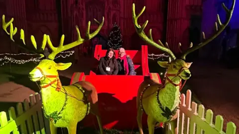 We Love Peterborough A red sleigh with two reindeers with golden decorations on them placed in front of the Cathedral, with two women sitting in it.