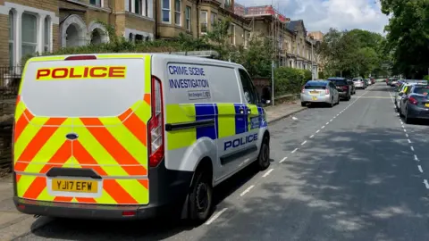 A Crime Scene Investigation police van parked on a street of terrace houses.
