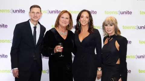 Teens Unite Fighting Cancer A man wearing a black suit, black tie and white shirt standing with three women in black dresses in front of a white backdrop with the logo of the charity Teens Unite Fighting Cancer printed on it.