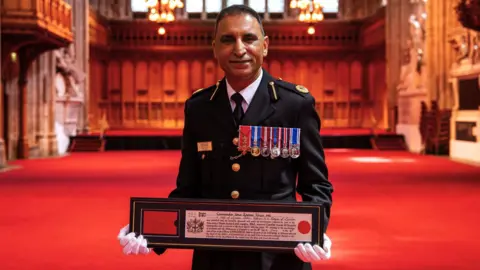 Umer Khan stands in traditional uniform with medals on display, holding up a certificate.
