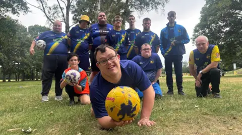 Naj Modak / BBC Group portrait of a community football team on a grassy park field surrounded by trees. Several participants wearing blue team kits pose together, while one participant in the foreground holds a yellow football toward the camera. Another participant kneels holding a white and purple football, with the rest of the team standing or kneeling behind. 