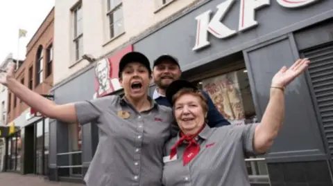 Somerset County Gazette Pauline in her KFC uniform of grey shirt and red neckerchief, being hugged by two other members of the team outside a KFC. 