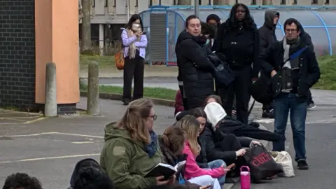 BBC/Simon Jones A queue of people outdoors some sat and some standing with some wearing masks