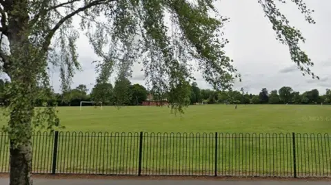 Google maps King George V playing fields in Haltemprice. The green playing fields are contained by a dark green fence. The trees are in leaf and there is a path running in front of the fence .
