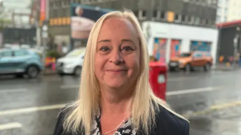 Karen McShane, a woman with shoulder-length, straight blonde hair, smiles at the camera. She is wearing a dark blazer and a black blouse patterned with flowers and a silver chain necklace. In the background is an out of focus street with parked cars.