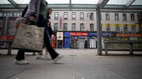 BBC Two people walking past a bus stop and shops in the centre of Glasgow. There is a row of different types of shops on the ground floor of a large building with rows of windows.