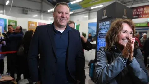 Getty Images CEO Mikael Peterson. He is tall with dark hair and smiling wearing a dark blue blazer, dark blue polo shirt and white t-shirt. A woman is standing next to him with dark hair wearing a clue coat. She is excitedly smiling holding her hands together.