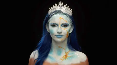Mia has dark blue hair. She is wearing a tiara and has a white and blue, frozen like, makeup look.