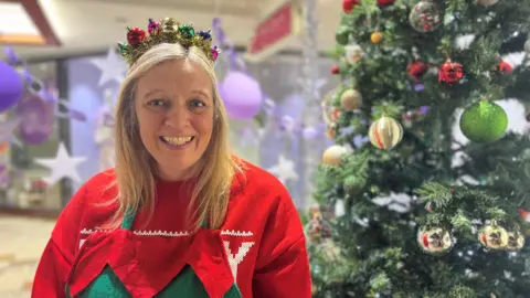 A woman in her fifties with blonde hair, she wears an elf outfit complete with a tinsel crown and is smiling. She stands next to a Christmas tree in the front window of the charity's base in a shopping centre.
